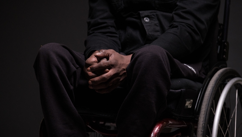 A person in a wheelchair in all black clothing and a dark skin tone clasping their hands together