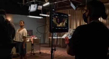 A masked crew member watches a monitor as several actors work on a scene
