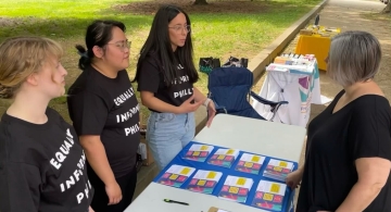 Three people wearing black t-shirts standing behind a table with printed cards as another person stands in front of the table
