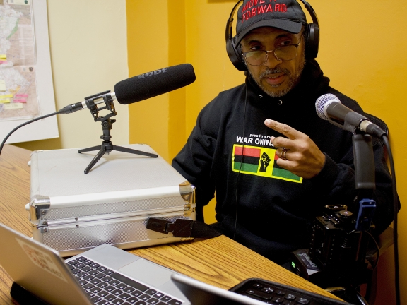 a man seated with two microphones pointing at him, making a point with one finger