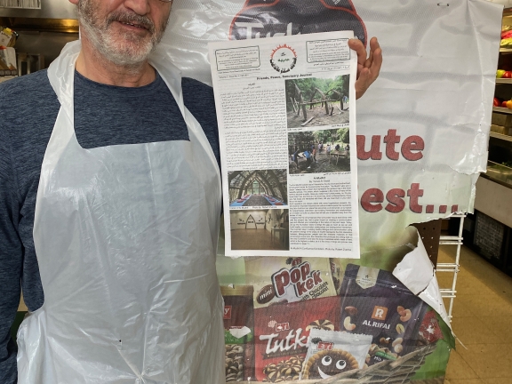 A person wearing a gray shirt, gray knit hat and a plastic apron holds up a newspaper