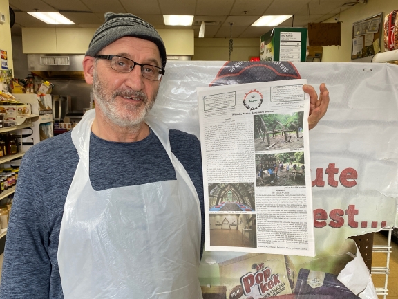 A close shot of a person wearing a gray shirt, gray knit hat and a plastic apron holds up a newspaper