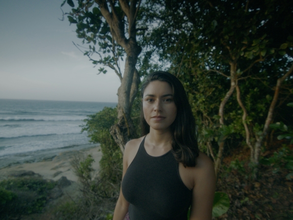 An olive toned person in a black halter top stands in front of trees on a beach