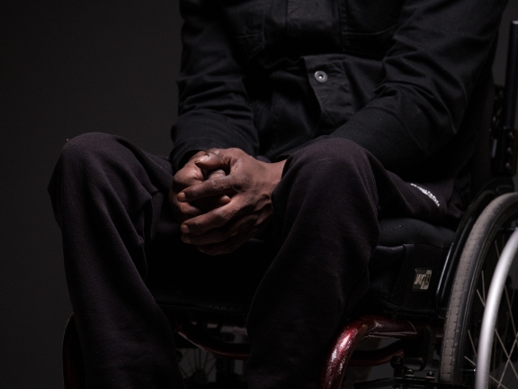 A person in a wheelchair in all black clothing and a dark skin tone clasping their hands together