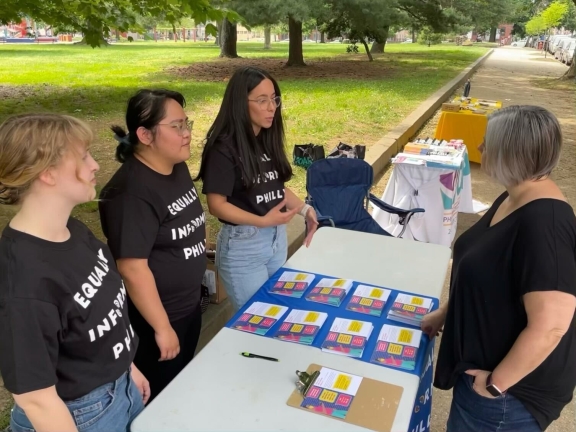 Three people wearing black t-shirts standing behind a table with printed cards as another person stands in front of the table