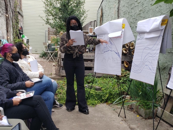 A person points to a drawing on a standing notepad in front of three sitting people