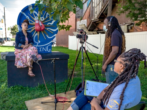 a woman sitting on a piece of public art while being interviewed by two people