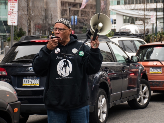 A man wearing glasses, a black hoodie and a kufi hat stands amidst traffic while speaking into a bullhorn
