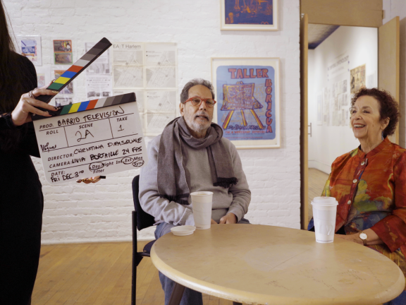 a behind the scenes film still of two people seated at a roundtable having a discussion and a person with a film slate standing to the left