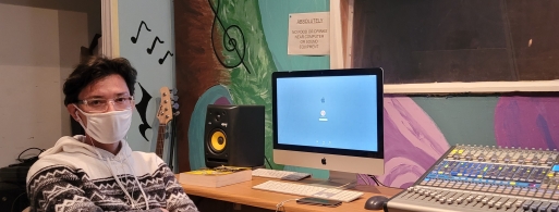 a masked person seated inside a music studio in front of a computer and audio equipment on a desk