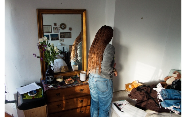 A person with brown braided hair, a gray top and blue jeans reaching into a corner past a dresser with a mirror on it