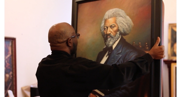 A bald person wearing glasses carrying a large portrait of Frederick Douglass in their hands