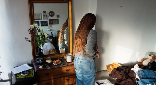 A person with brown braided hair, a gray top and blue jeans reaching into a corner past a dresser with a mirror on it