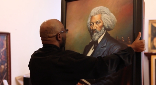 A bald person wearing glasses carrying a large portrait of Frederick Douglass in their hands