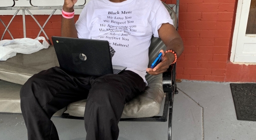 a man sitting on an outdoor couch with a laptop in his lap and phone in his hand giving a peace sign