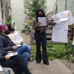 A person points to a drawing on a standing notepad in front of three sitting people