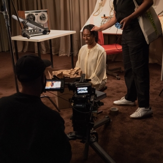 A film crew member adjusts the hair of a sitting person as another film crew member looks through a camera viewfinder