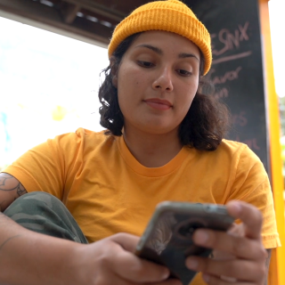 A light toned person with dark hair wearing a yellow knit hat, yellow t-shirt and green pants looks down at a phone